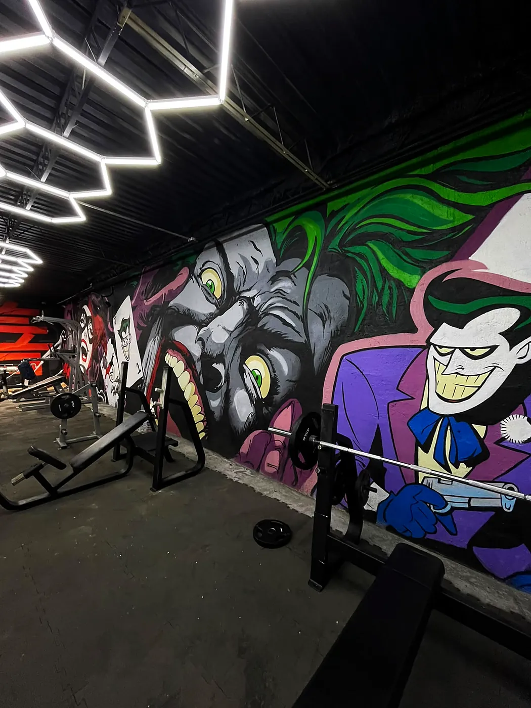 Piso de fuerza con bancos y barras frente al mural del Joker, bajo las lámparas hexagonales
